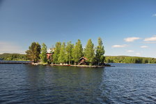 Eine kleine, bewaldete Insel mit Holzhäusern liegt in einem ruhigen See unter blauem Himmel.