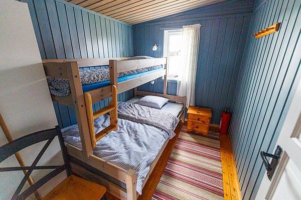Gemütliches Schlafzimmer in Hitra, Norwegen: Holzbett mit Etagen, blauer Holzwandanstrich, Fenster mit Vorhang, bunte Teppichstreifen. Ideale Unterkunft für Angelurlauber, die Komfort und Nähe zur Natur suchen. Optimal für Angelreisen gestaltet.