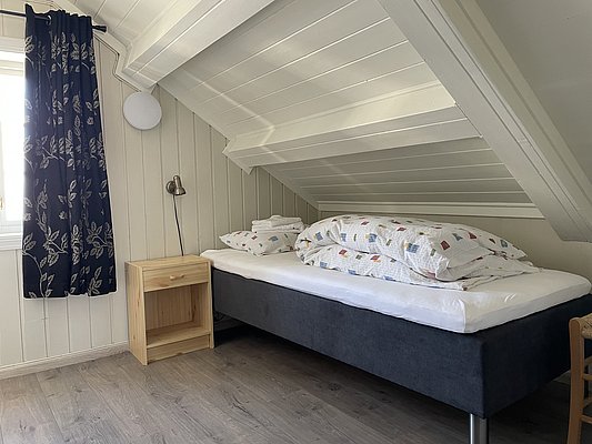 Gemütliches Schlafzimmer in Nord-Norwegen mit Einzelbett, Dachschräge, blumigen Vorhängen und Holzdetails, perfekt für einen erholsamen Angelurlaub. Helle, einladende Atmosphäre in typisch nordischer Einrichtung.