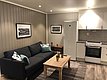 Gemütliche Ferienwohnung in Nord-Norwegen: Helles Wohnzimmer mit grauem Sofa, Holztisch und moderner Küche. Wandbilder, Pflanzen und dezente Beleuchtung schaffen eine einladende Atmosphäre. Ideal für Angelabenteuer und Entspannung in der atemberaubenden Natur.