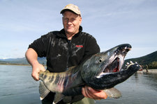 Ein Mann hält einen großen, gefangenen Lachs am Ufer eines Flusses in einer malerischen Landschaft.
