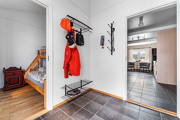 In einem gemütlichen Ferienhaus in Nord-Norwegen sieht man einen Flur mit Klinkerboden, angegliederter Wohnbereich mit großer Fensterfront, rustikalem Holzbett und Garderobe – ideal für Angelurlauber.