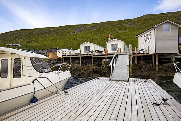 Hafen in Skarsvåg, Nord-Norwegen: Boote am Steg, umgeben von grünen Hügeln und Holzhäusern des Skarsvåg Turistsenters. Ideal für Angelreisen und atemberaubende Naturerlebnisse.