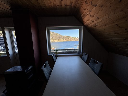 Gemütlicher Raum in Hitra, Norwegen, mit Holzdecke und Panoramafenster. Blick auf ruhigen Fjord und bewaldete Hügel. Ideal für Angler, die Entspannung und atemberaubende Natur erleben möchten. Perfekte Mischung aus Komfort und Abenteuer.