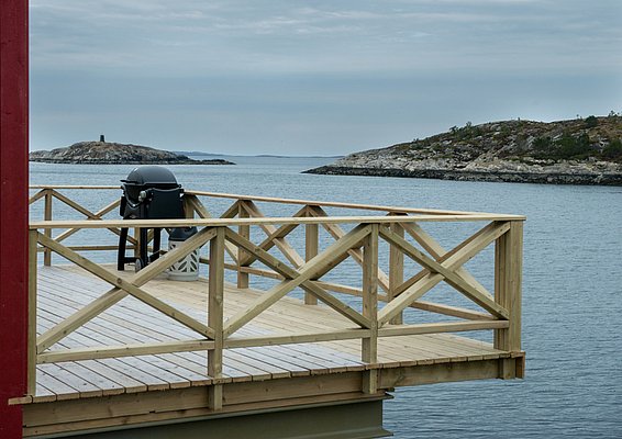 Eine Holzterrasse mit einem Grill bietet einen Blick auf eine ruhige Meereslandschaft mit felsigen Inseln im Hintergrund.