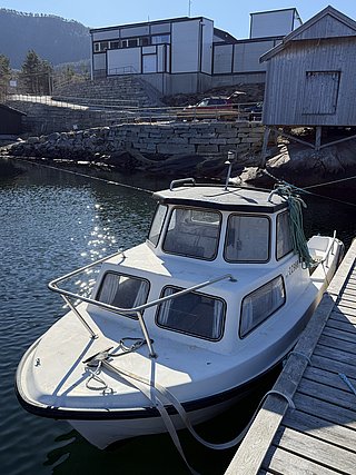 Ein weißes Fischerboot liegt in einem malerischen Hafen in Nord-Norwegen vor Anker. Holzhäuser und klare, reflektierende Wasseroberfläche schaffen eine ruhige, einladende Atmosphäre für Angelabenteuer.