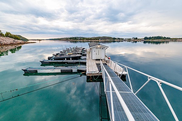 Ruhiger Hafen in Nordland, Norwegen: Anlegestelle mit Booten auf spiegelglattem Wasser, umgeben von sanften Hügeln. Wolkenverhangener Himmel reflektiert in der ruhigen See, perfekt für Angelreisen und entspannte Naturerlebnisse.