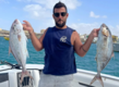 Ein Angler präsentiert stolz zwei gefangene Fische auf einem Boot in São Vicente, Kapverden. Im Hintergrund sind das Meer und die Küste zu sehen. Perfekte Szene für einen erfolgreichen Angelurlaub.