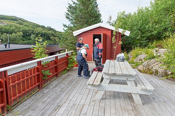 Eine Holzterrasse in der norwegischen Region Hitra, mit roten Gebäuden, grüner Umgebung und Picknicktisch. Ideal für Angelurlaub und Erholung. Ruhige, naturnahe Atmosphäre zum Verweilen in skandinavischer Landschaft.