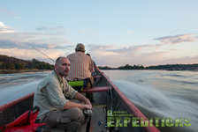Ein Boot gleitet über einen ruhigen Fluss in Südwest-Norwegen bei Sonnenuntergang. Zwei Angler fokussiert auf unberührte Landschaft. Ideal für Angelurlaub und Erkundung der nordischen Naturwunder.