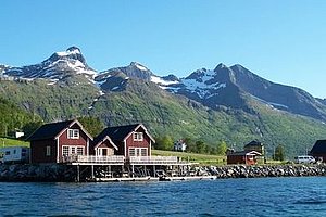 Rote Fischerhütten am Wasser in Nordland, Norwegen vor beeindruckender Bergkulisse. Sonnige Landschaft ideal für Angelurlaub und Meeresangeln. Perfekt für Abenteuerlustige, die Natur und Erholung suchen. Petri Heil – sorglos angeln weltweit!