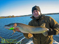 Ein Mann hält einen großen Hecht mit einem bunten Köder vor einer malerischen Seelandschaft.
