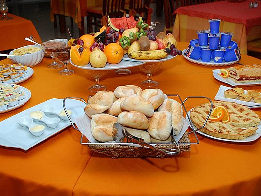 Auf einem Buffet sind verschiedene Frühstücksgerichte zu sehen, darunter Brot, Obst, Aufstriche, Joghurts und Kuchen auf einem Tisch mit orangefarbenem Tuch. Auf einem Buffet sind verschiedene Frühstücksgerichte zu sehen, darunter Brot, Obst, Aufstriche, Joghurts und Kuchen auf einem Tisch mit orangefarbenem Tuch.