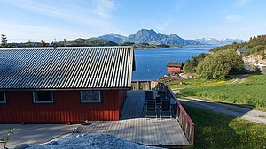 Idyllische Landschaft in Nordland, Norwegen: Rote Hütte mit Terrasse, Blick auf Fjord und majestätische Berge. Perfekt für Angelurlaub und Erholung in der Natur. Ideal für Abenteuerlustige und Ruhe Suchende.