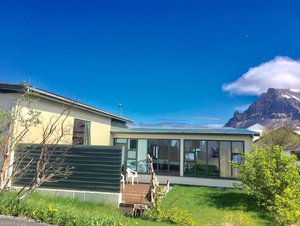 Ein einladendes Haus in Bolungarvik, Island, umgeben von üppigem Grün. Im Hintergrund erhebt sich majestätisch ein Berg unter strahlend blauem Himmel. Perfekter Ausgangspunkt für Natur- und Angelabenteuer.