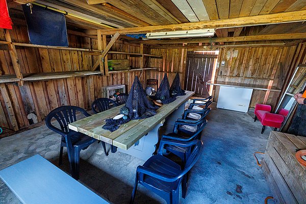 Gemütlicher Innenraum einer rustikalen Holzhütte in Hitra, Norwegen. Langer Tisch mit Netzen und Angelzubehör, umgeben von schwarzen Plastikstühlen. Perfekt für entspannte Angelpausen und gemeinsame Erlebnisse inmitten der Natur.