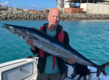 Ein Angler hält stolz einen großen Fisch (vermutlich ein Wahoo) auf einem Boot vor der malerischen Küste von São Vicente, Kapverden. Ideal für Angelreisen und unvergessliche Meeresabenteuer.