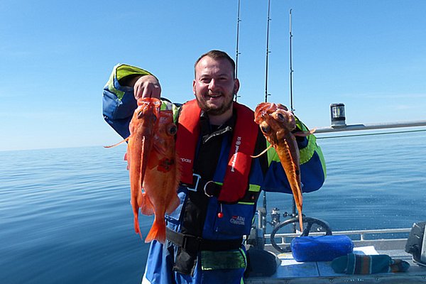 Ein Angler in Norwegen, Region Hitra, hält stolz mehrere frisch gefangene Fische auf einem Boot. Sonniger Himmel, ruhige See. Perfekter Angelurlaub für Meeresangler. Petri Heil – sorglos angeln weltweit!