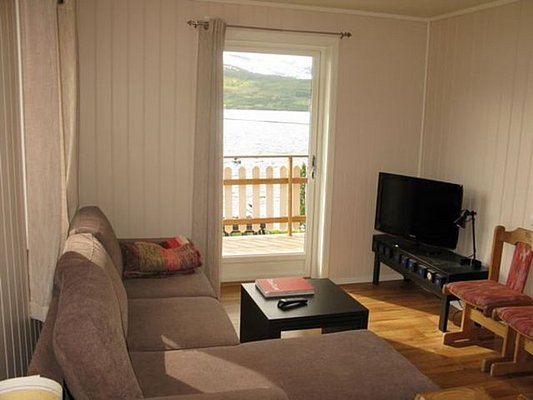 Gemütliches Wohnzimmer in Nord-Norwegen mit Sofa, Fernseher und Balkonblick auf fjordähnliche Landschaft. Ideal für Angelurlaub und Entspannung in ruhiger Umgebung. Perfekter Ausgangspunkt für Abenteuer und Erholung beim Angeln.