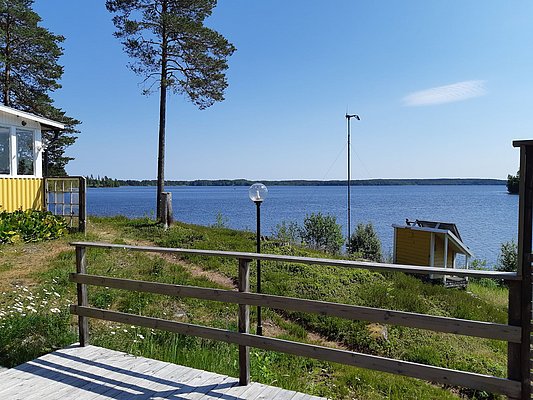 Ein sonniger Tag an einem ruhigen See mit einem Holzdeck, Bäumen und einem kleinen gelben Häuschen im Hintergrund.