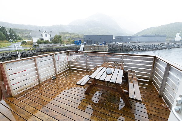 Nordland, Norwegen: Eine regennasse Terrasse mit Holztisch und Bänken, umgeben von malerischen Bergen und Küstenblick. Perfekt für Angelreisen mit eindrucksvoller Naturlandschaft und entspannten Momenten in rustikaler Umgebung.