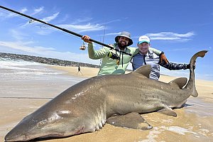 Zwei Angler in Nord-Norwegen posieren stolz am Strand mit einem großen gefangenen Hai. Ideal für Angelreisen und Abenteuerlust in einer atemberaubenden Landschaft. Perfekter Fangmoment für leidenschaftliche Meeresangler.