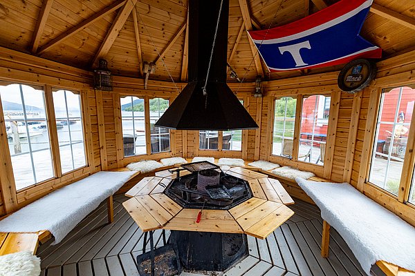 Ein gemütlicher, holzverkleideter Pavillon in Nord-Norwegen mit offenem Kamin, Sitzbänken und Blick auf die malerische Fjordlandschaft. Perfekt für einen entspannten Angelurlaub in eindrucksvoller Naturkulisse.