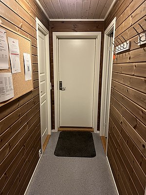Holzverkleideter Flur in einem norwegischen Haus, Nord-Norwegen. Zwei weiße Türen, grauer Teppich, Pinnwand mit Dokumenten. Gemütliche skandinavische Atmosphäre, perfekt für Angelurlaub in ruhiger Umgebung.