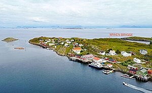 Luftaufnahme von Myken Brygge in Nordland, Norwegen: Malerisches Fischerdorf auf einer Insel, umgeben von ruhigem Meer. Farbenfrohe Häuser, Boote am Steg und weite Landschaften verstärken die idyllische Atmosphäre. Perfekter Ort für Angelreisen und Erholung.