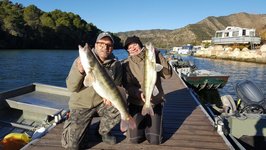 Zwei Personen posieren auf einem Steg an einem See mit zwei großen gefangenen Fischen.