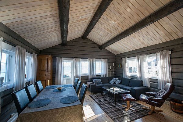 Gemütliches Wohnzimmer in einer Holzhütte in Nord-Norwegen. Die Einrichtung kombiniert moderne und rustikale Elemente mit einem großen Esstisch und Sofa. Fenster bieten Blick auf die Landschaft. Ideal für entspannende Angelreisen!