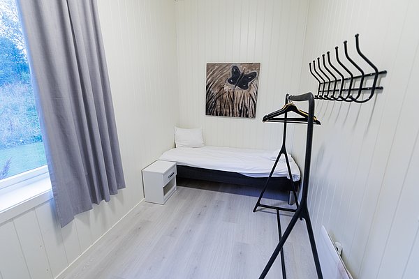 Kompaktes, helles Zimmer in Nordland, Norwegen, mit Einzelbett, schlichtem Bild an der Wand, Kleiderständer und Fenster mit Blick auf grünen Außenbereich. Ideal für entspannenden Angelurlaub.