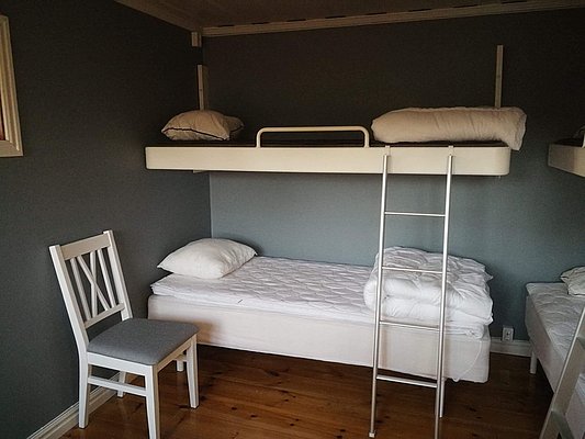 In einem gemütlichen Raum mit Holzboden befinden sich ein Etagenbett mit Leiter, zwei Einzelbetten und ein weißer Stuhl vor einer grauen Wand.