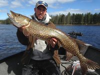 Eine Person hält einen großen Hecht in einem Boot auf einem See, umgeben von Wald und blauem Himmel.