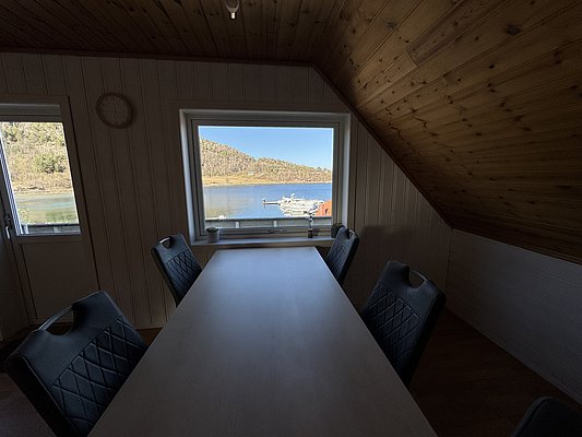 Ein heller Raum mit Holzwänden bietet einen malerischen Ausblick auf norwegische Küstenlandschaft und Boote. Perfekt für Angelreisende, die Ruhe und Natur genießen möchten. Ideal für entspannte Angelurlaube in Hitra, Norwegen.