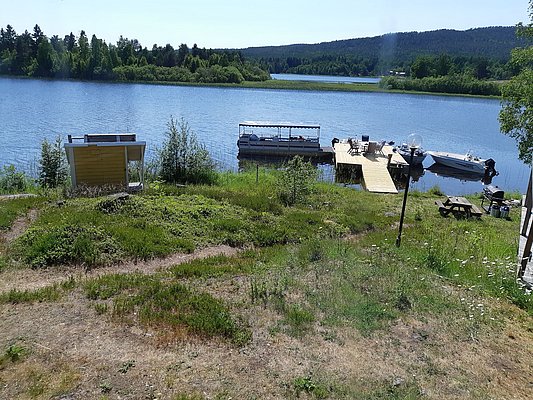 Ein ruhiger See mit einem Holzsteg, an dem zwei Boote festgemacht sind, umgeben von grüner Landschaft und einem blauen Himmel.