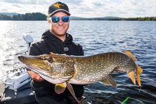 Ein Mann auf einem Boot hält einen großen Hecht hoch, während im Hintergrund ein See und bewaldete Ufer zu sehen sind.