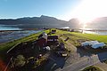 Luftaufnahme einer idyllischen Farm in Nord-Norwegen mit roten Gebäuden und weiten Feldern, umrahmt von majestätischen Bergen und einem glitzernden Fjord bei strahlendem Sonnenschein. Perfekt für Angelreisen und Naturliebhaber.