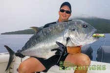 Ein glücklicher Angler hält einen riesigen GT-Riesen-Trevally an Bord eines Boots in Tansania. Traumhafte Kulisse mit bewölktem Himmel und grüner Insel im Hintergrund. Perfekter Angelurlaub für Abenteurer.