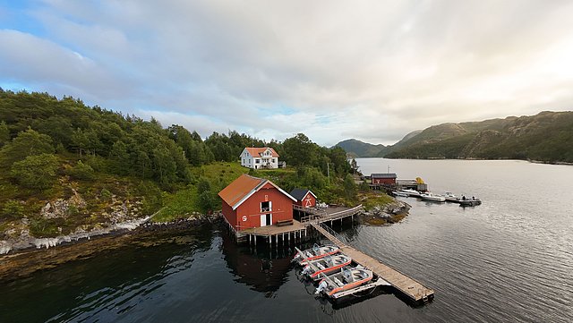 Küstenlandschaft in Hitra, Norwegen: Rote Fischerhütten mit Bootssteg am ruhigen Fjord, umgeben von grünen Wäldern und Bergen. Perfekter Angelurlaubsort für Naturliebhaber und Abenteurer. Ideal für Angelreisen und maritime Erkundungen.