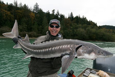 Eine Person hält einen großen Stör vor einer bewaldeten Flusslandschaft hoch.