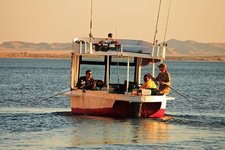 Ein kleines Boot mit drei Menschen beim Angeln fährt auf einem ruhigen Gewässer, im Hintergrund sind sandige Hügel zu sehen.