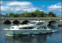 Ein modernes Hausboot fährt auf einem Fluss vor einer Steinbrücke bei sonnigem Wetter.