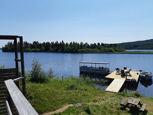 Bild eines sonnigen Sees mit einem hölzernen Anlegesteg, auf dem Tische und Stühle stehen, flankiert von einem Boot und umgeben von grüner Landschaft.