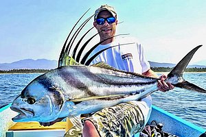 Ein Angler hält einen großen Hahnenfisch auf einem Boot vor einer malerischen Landschaft aus Wasser und Bergen.