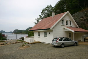 Gemütliches weißes Ferienhaus in Süd-Norwegen, umgeben von Waldlandschaft. Ein Auto steht daneben. Im Hintergrund ruhiger Fjord, ideal für Angelurlaub und Naturerlebnis. Perfekte Unterkunft für Abenteurer und Erholungssuchende.