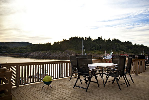 Holzveranda mit Sitzmöglichkeiten bietet Blick auf die norwegische Küste in Nordland. Sonnenuntergang über Bäumen und Wasser. Ideal für Angelurlaub und Entspannung. Perfekte Ausgangslage für Meeresangeln und Naturerlebnisse.