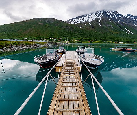 Angelboote an einem hölzernen Steg, eingebettet in die beeindruckende Landschaft Nord-Norwegens mit schneebedeckten Bergen. Ideal für Angelreisen und unvergessliche Abenteuer. Petri Heil – sorglos angeln weltweit.