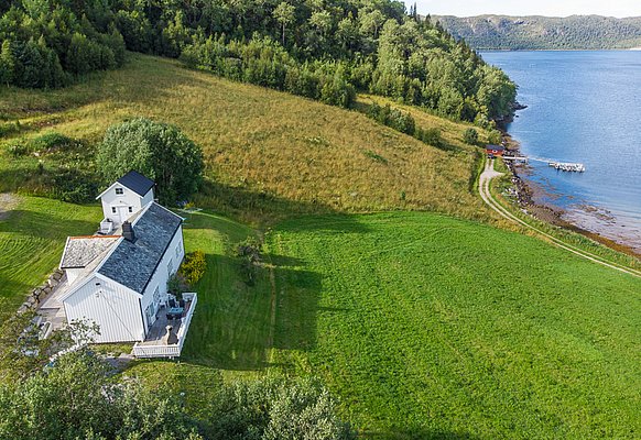 Ländliche Idylle auf Hitra, Norwegen: Weißes Haus umgeben von grünen Feldern und dichten Wäldern, direkt an der malerischen Küste mit Bootsanleger. Perfekter Ort für einen erholsamen Angelurlaub in beeindruckender Natur.