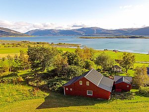 Grüne Landschaft mit rotem Haus am Ufer eines Fjords in Nordland, Norwegen. Malerische Aussicht auf ruhiges Wasser und Berge am Horizont. Ideal für naturnahen Angelurlaub.